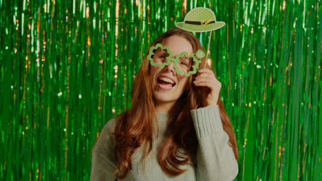 Woman-Celebrating-St-Patrick's-Day-Standing-In-Front-Of-Green-Tinsel-Curtain-Wearing-Prop-Shamrock-Shaped-Glasses-1